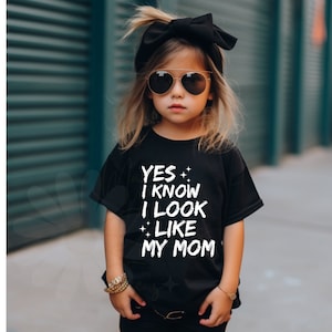 May include: A young girl wearing a black t-shirt with the text "YES I KNOW I LOOK LIKE MY MOM" in white. She is wearing sunglasses and a black headband.