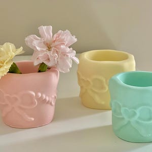 May include: Three pastel-colored ceramic pots with a bow and heart design. The pots are pink, yellow, and mint green. The pink pot holds a pink flower, and the yellow pot is empty. The pots are on a white surface.