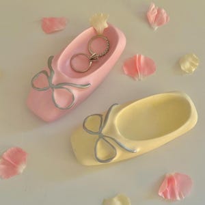 May include: Two ballet slipper-shaped ring holders, one pink and one yellow, each with a silver bow detail. One pink holder contains two rings. Scattered around are pink and cream-colored flower petals.