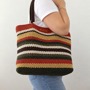 May include: A rectangular crochet tote bag featuring horizontal stripes in shades of dark green, mustard yellow, rust orange, brown, and cream. The bag has dark brown crochet handles attached with gold-toned rings and a small brown leather label.