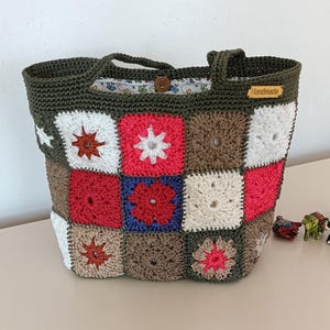 May include: A green crocheted tote bag with a patchwork design of colorful squares featuring different flower patterns. The bag has a brown leather tag that says "handmade".