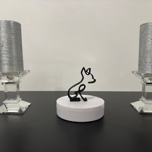 Peut inclure: Une sculpture en métal noir d'un chien assis sur une base ronde blanche. La sculpture est sur une surface noire avec deux chandeliers argentés de chaque côté.