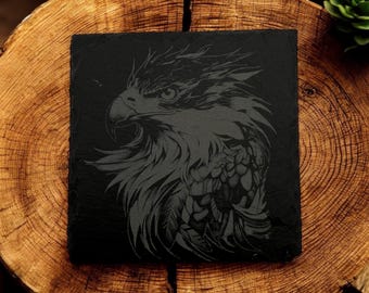 Puede incluir: Un posavasos de pizarra negra con un grabado detallado de la cabeza de un águila. Las plumas del águila están talladas de forma intrincada y su ojo es prominente.