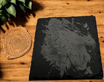 Puede incluir: Un posavasos de pizarra negra con un grabado detallado de la cabeza de un águila con plumas y elementos florales. El pico del águila está abierto y el grabado es en tono gris.