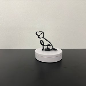 May include: A black wire sculpture of a dog sitting on a white pedestal. The sculpture is a simple line drawing of a dog.