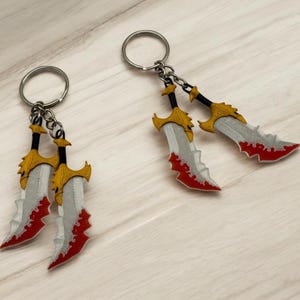 Könnte beinhalten: Schlüsselanhänger mit Miniatur-Doppelklingen-Schwertern. Die Klingen sind silberfarben mit roten Akzenten, die Griffe gold und schwarz. Jeder Schlüsselanhänger hat einen silbernen Ring zum Befestigen.