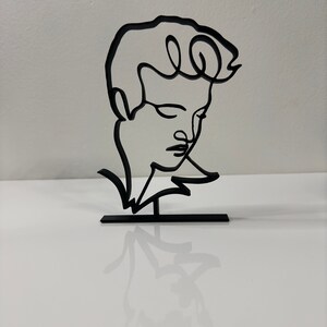 May include: Black metal line art sculpture of a person's head and shoulders. The sculpture is on a small black metal base.