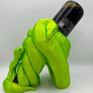 Könnte beinhalten: Ein leuchtend grüner, schlangenförmiger Weinflaschenhalter. Der Halter ist aus Kunststoff und hat ein realistisches Schlangendesign. Eine Weinflasche ist teilweise in den Halter gesteckt.