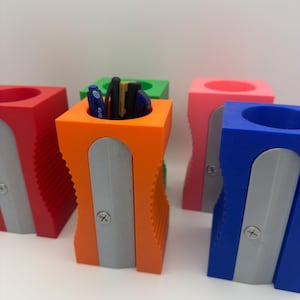 Könnte beinhalten: Fünf bunte Bleistiftspitzer mit integriertem Stifthalter. Die Spitzer sind rot, orange, grün, rosa und blau. Jeder Spitzer hat ein silbernes Messer und eine Schraube.