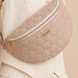 Könnte beinhalten: Eine beige gesteppte Bauchtasche mit sechseckigem Muster, ausgestattet mit einem goldenen Reißverschluss mit zwei runden Zippern und einem rechteckigen Aufnäher mit der Aufschrift Thalainy's auf der Vorderseite. Die Tasche hat einen verstellbaren Riemen und weiße Kanten.