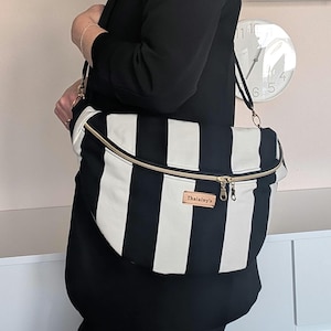 Könnte beinhalten: Eine halbmondförmige Umhängetasche mit einem markanten vertikalen Streifenmuster in Schwarz und Weiß. Die Tasche verfügt über einen goldfarbenen Reißverschluss und ein rechteckiges braunes Etikett mit dem Text Thalainy's auf der Vorderseite.