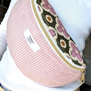 May include: A pink corduroy fanny pack with a patterned flap featuring a floral design in green, purple, and brown. The bag has a gold zipper and a white label that reads "Thalainys".