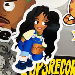 May include: A cartoon sticker of a woman with long black hair wearing a blue jumpsuit, a yellow shirt, and white trainers. The sticker is on a black and yellow background with the text "KOPS RECORD" in yellow.