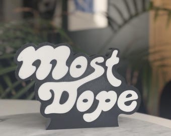 May include: A black and white sign with the words "Most Dope" in a retro font. The sign is on a marble surface, with a blurred background of greenery and a window.
