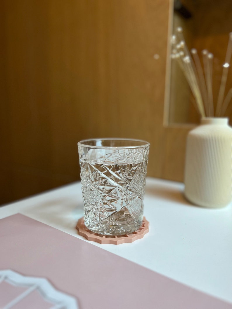May include: A clear glass with a cut-glass design filled with water sits on a pink coaster. The coaster has a decorative, geometric design.