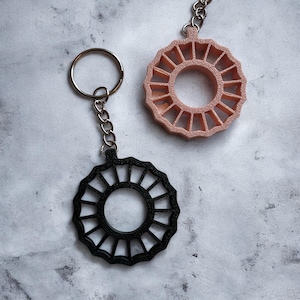 Puede incluir: Dos llaveros con un diseño circular. Uno es negro, el otro es rosa claro. Ambos tienen un anillo y una cadena para sujetar a las llaves. El diseño presenta líneas radiales alrededor de un círculo central.