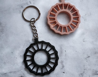 May include: Two keyrings with a circular design. One is black, the other is a light pink. Both have a ring and chain for attaching to keys. The design features radial lines around a central circle.
