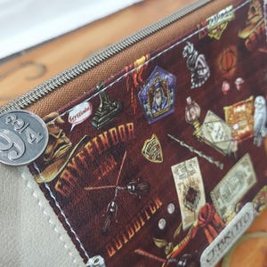 May include: A brown zippered pouch with a white leather trim. The pouch is decorated with a colorful print featuring Harry Potter themed images, including the Gryffindor crest, a Hogwarts acceptance letter, and a golden snitch. The pouch has a metal zipper pull with the number "9 3/4" on it.