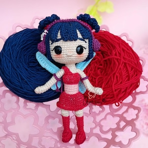 Op de afbeelding: Handgemaakte gehaakte pop met een feeënontwerp. De pop heeft een rode jurk, laarzen en vleugels, met blauw haar en een hoofdband. De pop staat op een roze bloemenbak met blauwe en rode bollen wol op de achtergrond.