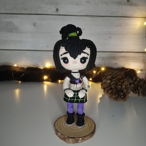 Puede incluir: Una muñeca de crochet con cabello negro, una falda de cuadros verdes y negros y botas moradas. La muñeca está de pie sobre una base de madera.