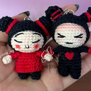May include: Two handmade crochet dolls, one in red and one in black, each with a unique design. The red doll has a red outfit and the black doll has a heart on its chest. Both dolls have black hair and are holding a small object.