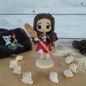 May include: A crocheted doll of Moana from the Disney movie Moana, holding a wooden paddle. The doll is wearing a red and white dress and has brown hair. The doll is standing on a wooden base.