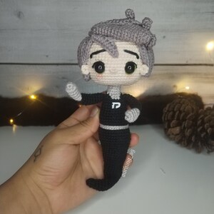 Pode incluir: Um brinquedo de pelúcia de crochê de um personagem com cabelo cinza, uma camisa preta com uma letra "P" branca na frente e uma cauda preta. O brinquedo está sendo segurado em uma mão.