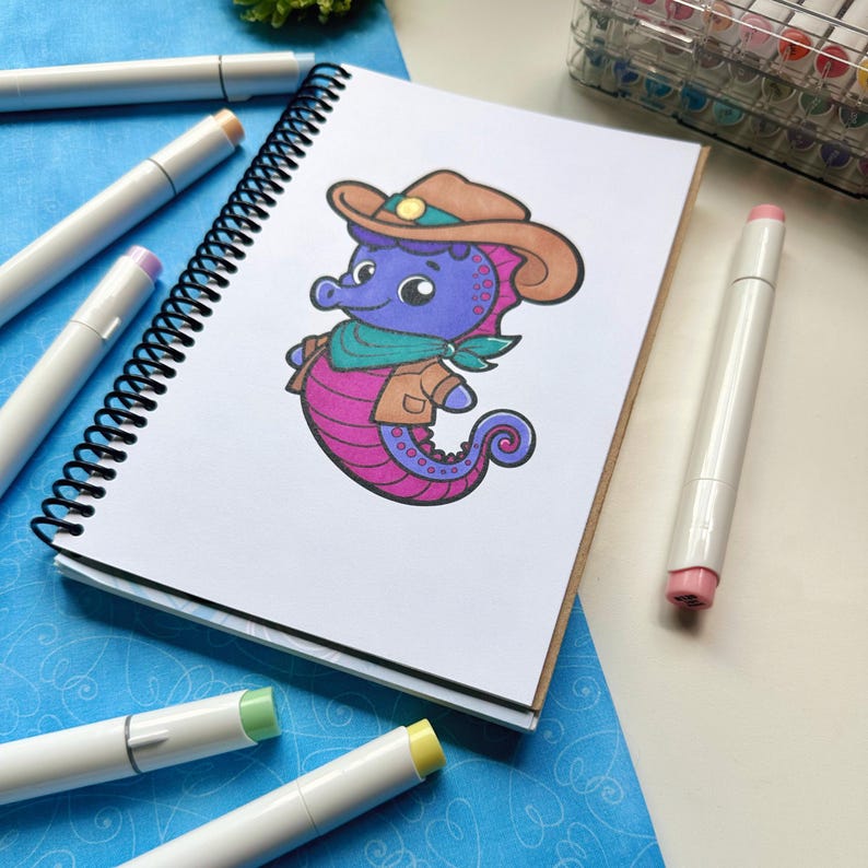 May include: A spiral-bound sketchbook with a colorful illustration of a seahorse wearing a cowboy hat, jacket, and scarf. The seahorse is purple and pink. Several white art markers with colored tips are scattered around the sketchbook on a blue patterned surface.