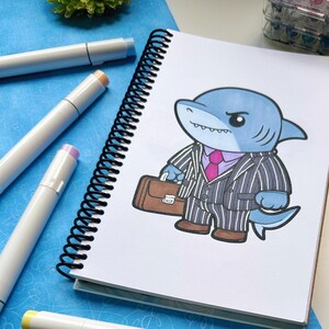 May include: A spiral-bound notebook with a cartoon shark character in a pinstripe suit, holding a briefcase. The shark is blue with a pink tie. Several art markers in various colors are scattered around the notebook.