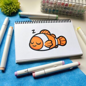 May include: A white spiral-bound sketchbook with a cartoon illustration of a sleeping orange and white clownfish. The word "Z" is above the fish. Several white art markers with colored tips surround the sketchbook on a blue surface.