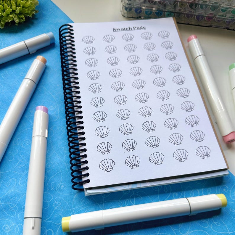 May include: A spiral-bound notebook with a seashell pattern on the pages, labeled "Swatch Page." Several white markers with colored tips, including pink, yellow, and blue, are scattered around the notebook on a blue surface.