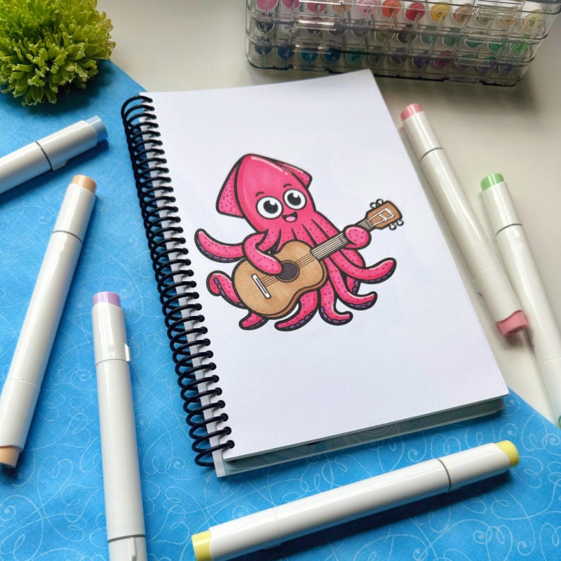 May include: A spiral-bound sketchbook featuring a pink cartoon squid playing a brown acoustic guitar. The squid has large eyes and is surrounded by white markers with colored tips. The background is a blue patterned surface.