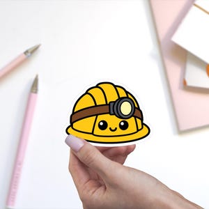 May include: A yellow hard hat sticker featuring a brown strap and a small headlamp attached to the front. The hard hat has a cartoon face with two black eyes and a small smiling mouth, rendered in a bold, graphic style with thick black outlines.