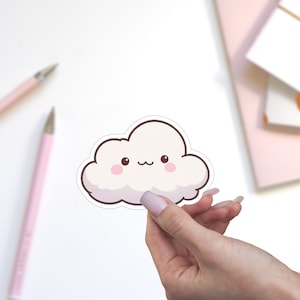 May include: A white cloud sticker with a cute cartoon face. The cloud has pink cheeks and a small smile. The sticker is being held by a hand.