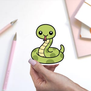 May include: A die-cut sticker featuring a cartoon snake with a light green body, darker green spots, and a cream-colored underbelly. The snake has large black eyes, a small smile, and a pink forked tongue protruding from its mouth.