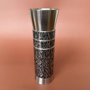 May include: A silver-colored vase with a textured, patterned design around the middle. The top flares out, and the base is a solid silver color. The vase is cylindrical and stands on a solid base.