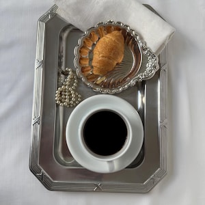 Op de afbeelding: Een zilveren dienblad met een witte kop zwarte koffie, een klein zilveren schaaltje met een croissant en een witte servet. Een parelketting is op het dienblad gedrapeerd. Het dienblad heeft een geometrisch ontwerp.