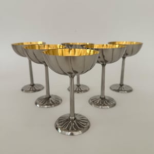 Op de afbeelding: Een set van zes zilverkleurige metalen coupes gerangschikt in een driehoekige formatie. Elk glas heeft een geribbelde, gefluteerde kom met een gepolijste goudkleurige binnenkant, een slanke steel en een bijpassende geribbelde ronde voet.