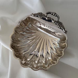 May include: A polished silver-toned metal dish shaped like a scallop shell. The dish features a scalloped edge and a decorative, ornate handle at the top with intricate scrollwork and embossed detailing. The surface is reflective and smooth.