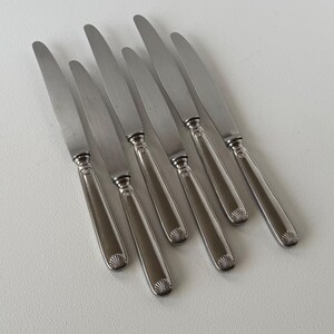 Pode incluir: Um conjunto de seis facas de jantar em aço inoxidável. Cada faca tem uma lâmina longa e fina e um cabo com um design decorativo de concha na base. As facas estão dispostas sobre uma superfície branca.
