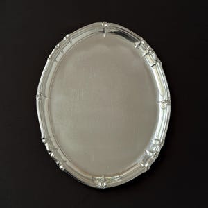 Könnte beinhalten: Ein ovales, silberfarbenes Serviertablett mit einer glatten, reflektierenden Oberfläche und einem dekorativen, gewellten Rand, der durch erhabene, sich wiederholende vertikale Segmente entlang der gesamten Kante eine strukturierte Optik erhält.