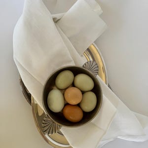 Puede incluir: Un pequeño cuenco plateado antiguo lleno de seis huevos, algunos verde pálido y otros naranja, descansa sobre una bandeja plateada. Una servilleta de lino blanco está drapeada sobre la bandeja, creando una presentación elegante. El fondo es blanco.