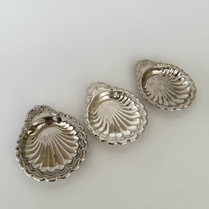 Op de afbeelding: Drie zilverkleurige, schelpvormige schalen. Elke schaal heeft een geschulpte rand en een gedetailleerde handgreep. De binnenkant van elke schaal heeft een geribbeld ontwerp. De schalen zijn op een wit oppervlak geplaatst.