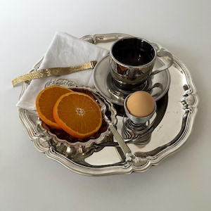 Può includere: Un vassoio d'argento con una tazza di caffè, un uovo alla coque e due fette d'arancia. Un braccialetto d'oro e un tovagliolo bianco sono anche sul vassoio. Il vassoio ha un bordo decorativo smerlato. Le fette d'arancia misurano circa 7 cm di diametro.