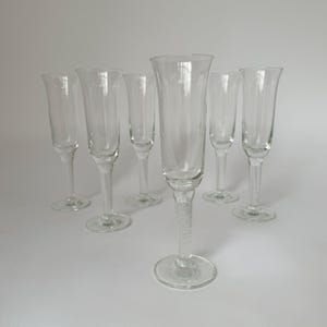 Può includere: Un set di sei flûte da champagne in vetro trasparente con coppe slanciate e steli testurizzati. Ogni flûte presenta un'apertura alta e stretta e una base circolare, disposte in gruppo con un bicchiere posizionato in primo piano.