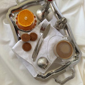 Puede incluir: Una bandeja plateada con media naranja, dos gofres pequeños, cubiertos, un vaso de café y un plato en forma de concha. Una vela roja está en un candelabro plateado. La bandeja está sobre un paño blanco.