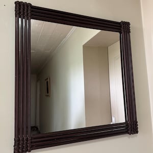 Puede incluir: Un espejo de pared cuadrado con un marco de color marrón oscuro construido con múltiples varillas paralelas. Las esquinas del marco están reforzadas con un detalle trenzado tipo celosía que une las varillas, creando un borde texturizado.