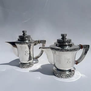 May include: Two silver-toned metal teapots featuring ornate embossed floral detailing around the base, lid, and handle. Each teapot displays a central engraved monogram letter E and includes a matching lid with a tiered, circular finial top.
