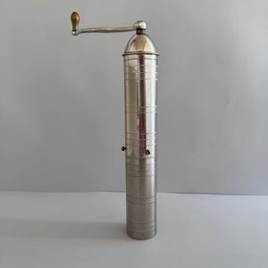 May include: A tall, cylindrical silver-toned metal spice grinder featuring a polished finish with horizontal engraved bands. The top includes a silver handle with a small, rounded brass-colored knob for manual operation and a dome-shaped cap.