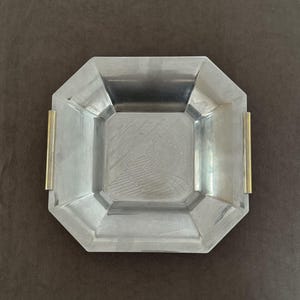 Puede incluir: Una bandeja cuadrada de metal plateado con bordes biselados y un patrón geométrico grabado en el centro. La bandeja cuenta con dos asas verticales de metal dorado fijadas en los lados opuestos, contrastando con el acabado plateado.